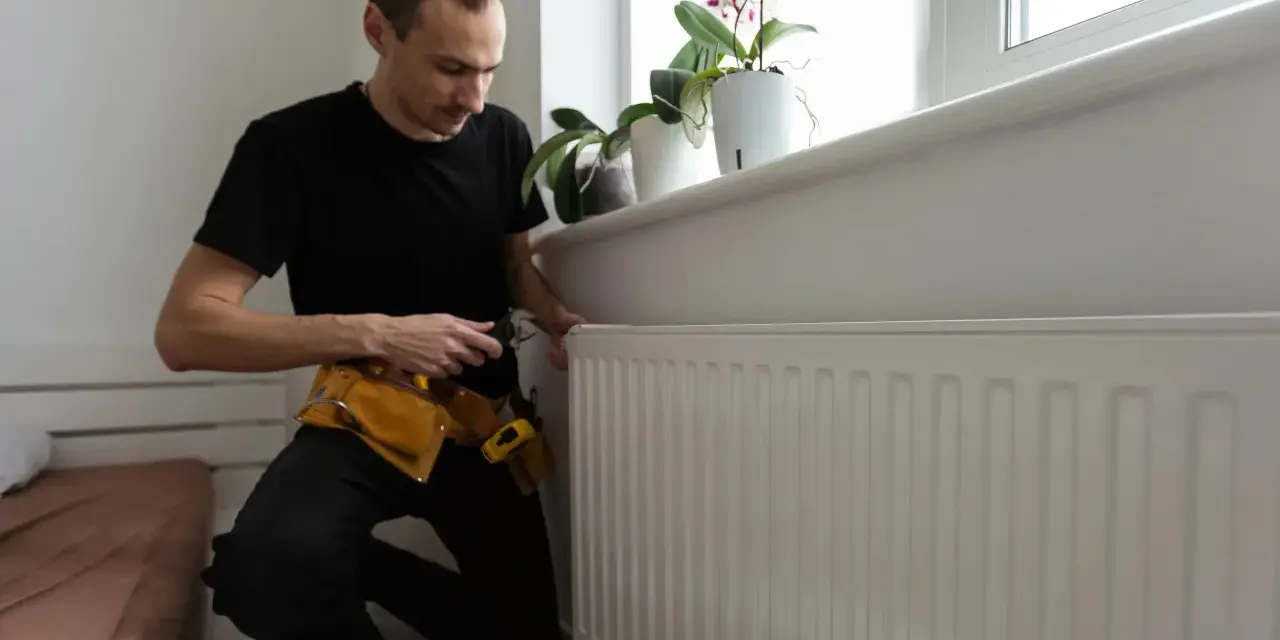 A man in a tool belt inspects a white radiator, checking for any signs of a cracked radiator before winter sets in.