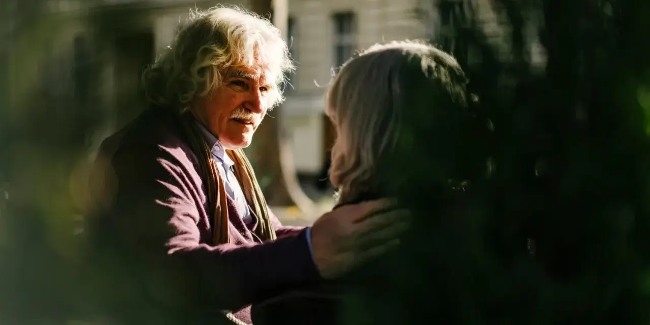 A man with white, curly hair and a mustache gently places his hand on another person's shoulder, offering a comforting response. This scene illustrates react vs respond examples.