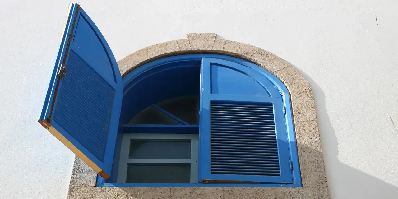 Finestre aperte o chiuse d'estate, con persiane blu su muro bianco. La luce del sole crea ombre nette.