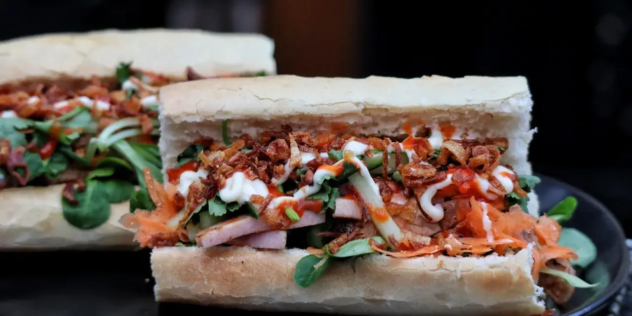 Zwei appetitliche Banh Mi Sandwiches mit Fleisch, Gemüse, knusprigen Zwiebeln und Saucen auf einem Teller.