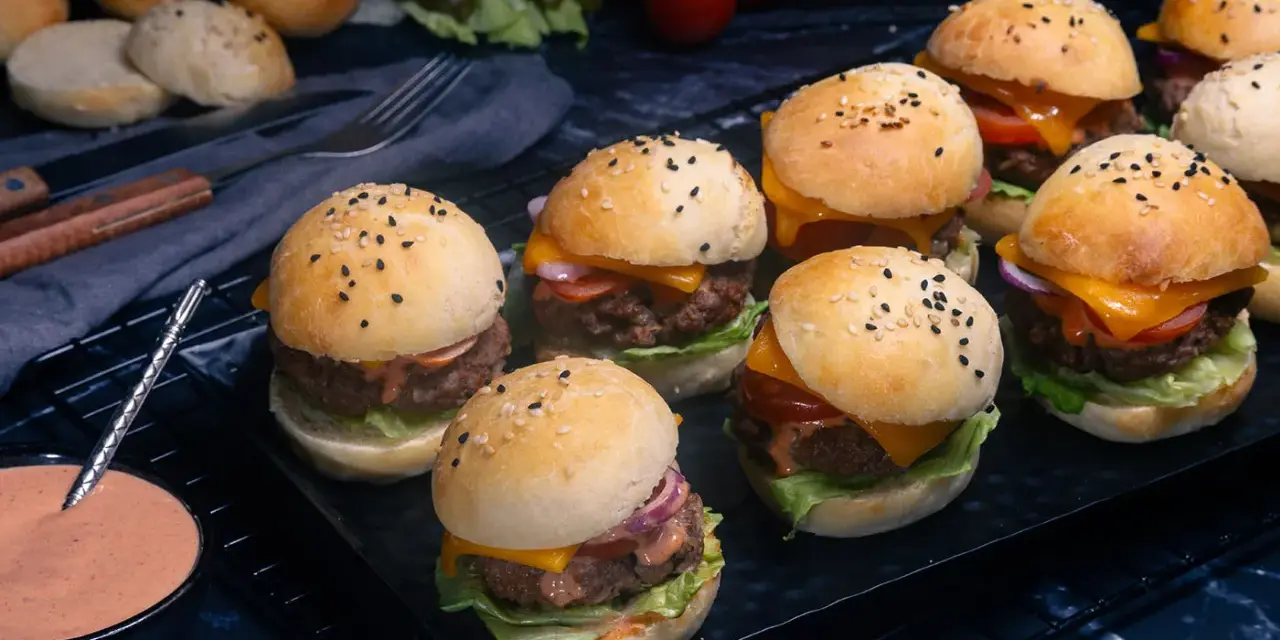 Mehrere mini burger mit Käse, Salat und Tomaten auf einem Rost. Daneben eine Schale mit Sauce.