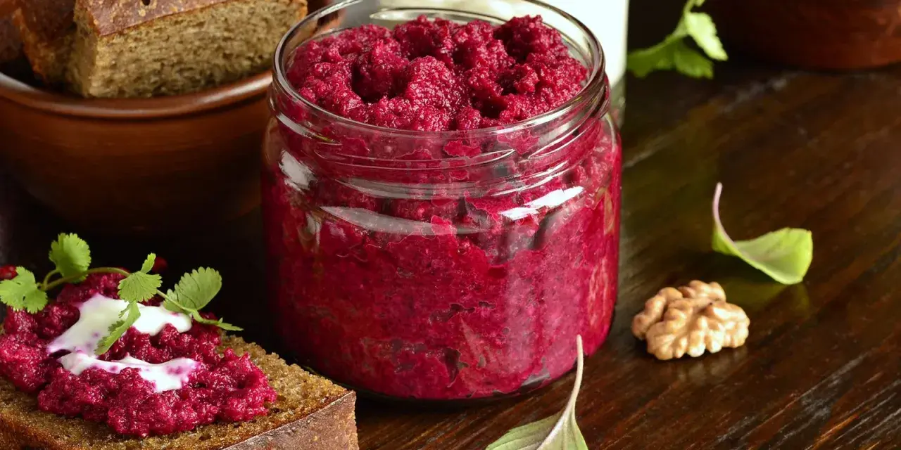Glas mit leuchtend rotem Rote Bete Pesto, daneben Brot mit Pesto und Joghurt. Walnuss und Kräuter garnieren die Szene.