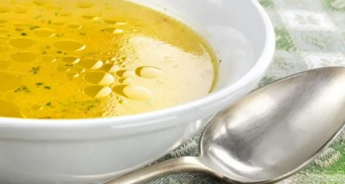Una ciotola bianca piena di brodo di carne dorato, con goccioline d'olio in superficie. Un cucchiaio d'argento è appoggiato accanto.