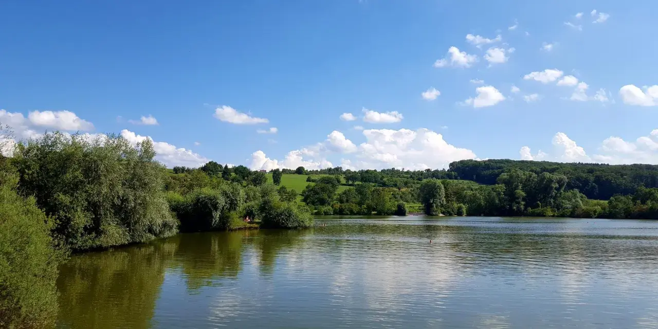 Ein sonniger Tag am Badesee Esslingen. Grüne Hügel, Bäume und ein blauer Himmel mit weißen Wolken spiegeln sich im ruhigen Wasser.