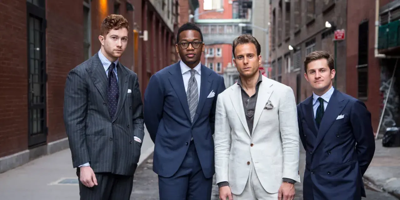 Quattro uomini in abiti eleganti, con un impeccabile bavero giacca, posano in un vicolo urbano.