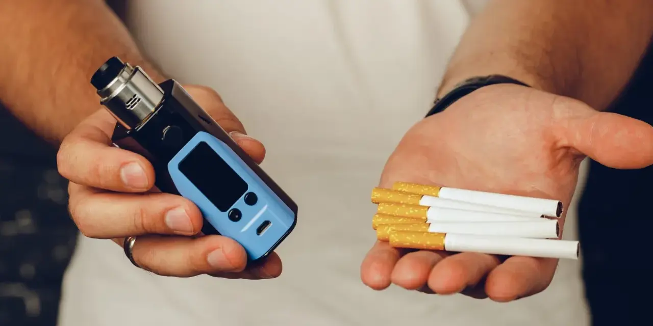 Man holds a vape in one hand and cigarettes in the other, posing the question: que es mejor el vaper o el tabaco.