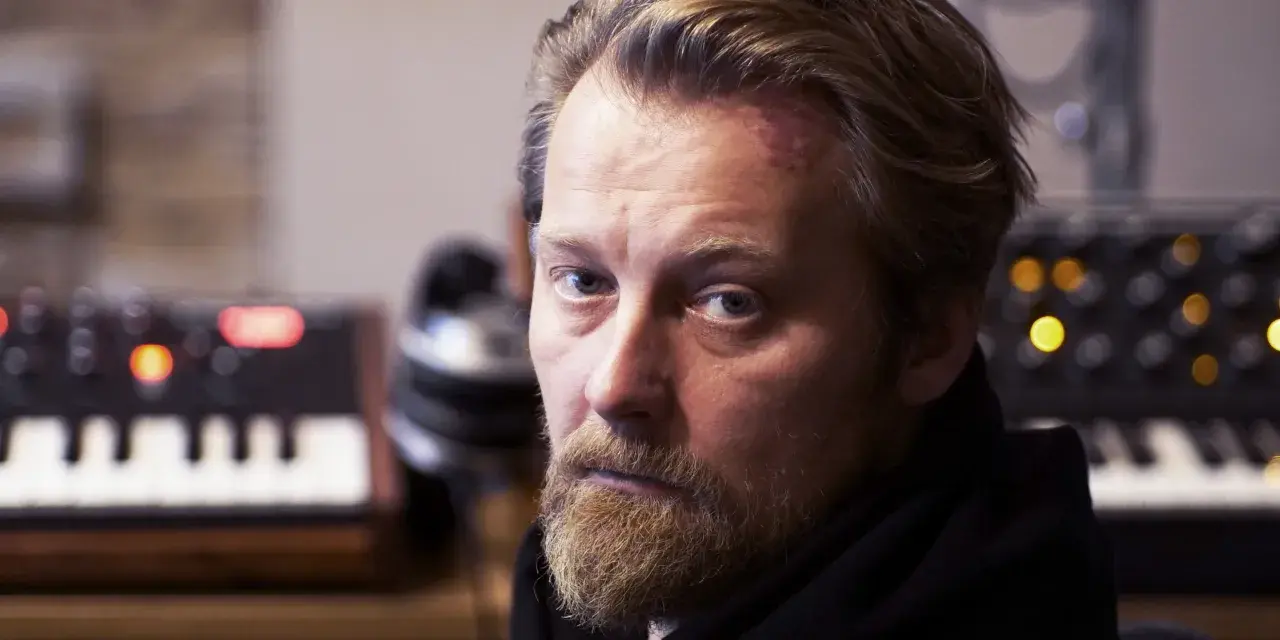 Bartek Dziedzic, z zarostem i lekko zaczerwienionym czołem, siedzi przed syntezatorami.