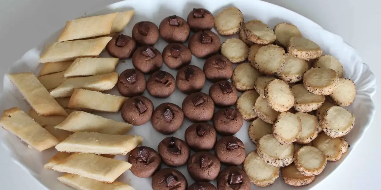 En vit oval bricka fylld med tre sorters hembakade småkakor: rombiska, chokladpraliner och runda med strössel. Perfekt för fika!