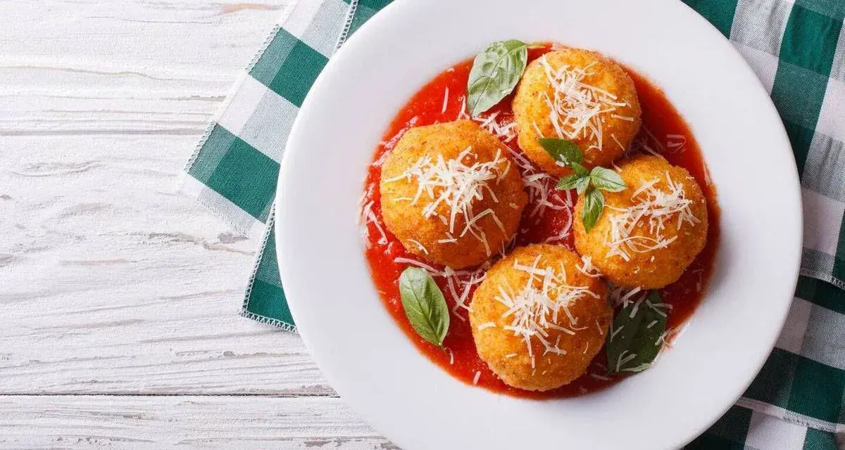 Quattro arancini di riso dorati, cosparsi di parmigiano e serviti su un letto di salsa di pomodoro, guarniti con basilico fresco.