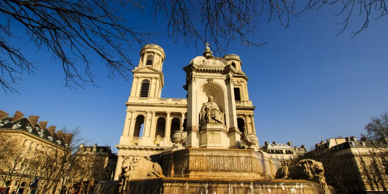 Fuente monumental frente a la iglesia de Saint Sulpice, con leones de piedra y una estatua central bajo un cielo azul claro.