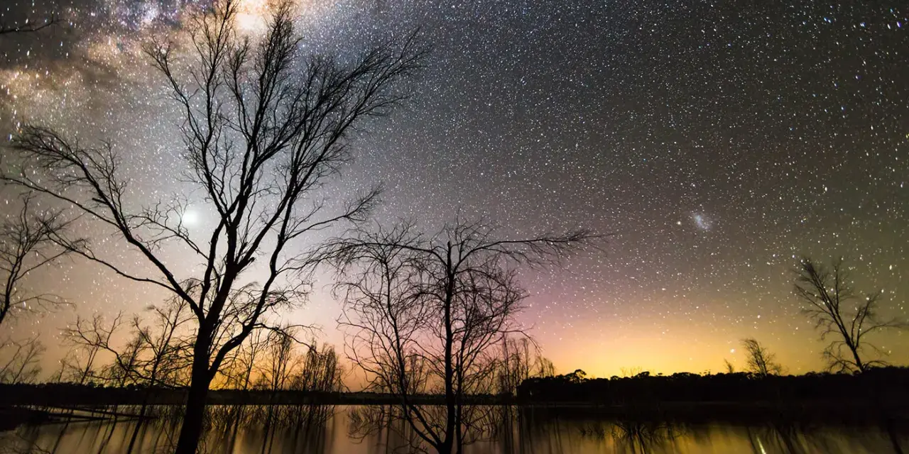 Canon EOS 2000D photo de nuit : une voie lactée spectaculaire au-dessus d'un lac reflétant les arbres dénudés et un ciel orangé.