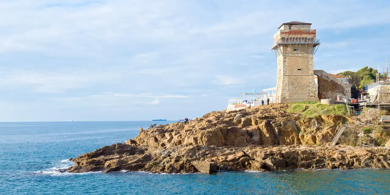 Torre medievale e mare blu a Calafuria, con scogli rocciosi e cielo nuvoloso.