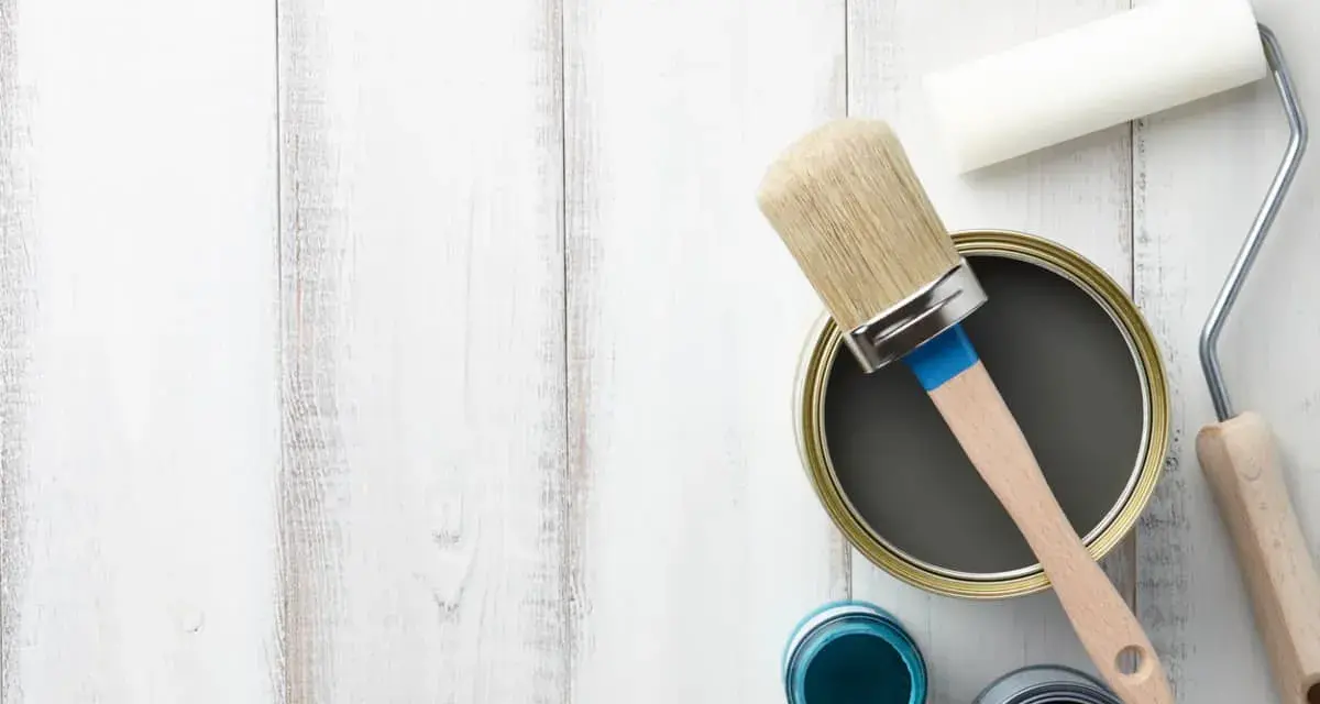 Herramientas para pintar mesitas de noche: lata de pintura gris, pincel, rodillo y botes de pintura azul y negra sobre fondo de madera blanca.