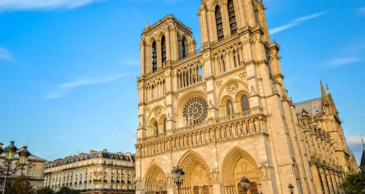 La majestueuse façade de Notre Dame de Paris se dresse sous un ciel bleu, ornée de détails gothiques. Des passants flânent sur la place.