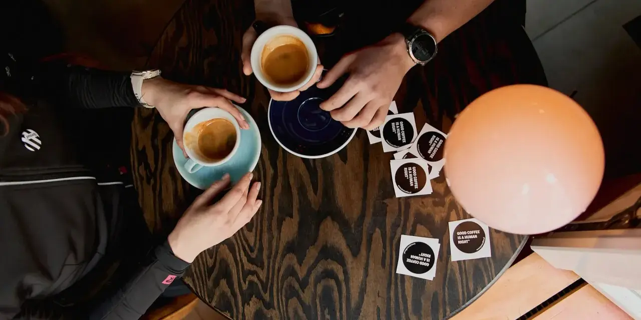 Dwie osoby przy stoliku piją kawę. Jedna z nich trzyma filiżankę, druga sięga po nią. Na stole leżą naklejki z napisem "GOOD COFFEE IS A HUMAN RIGHT". To idealny moment na rozmowę o autofagii i kawie.