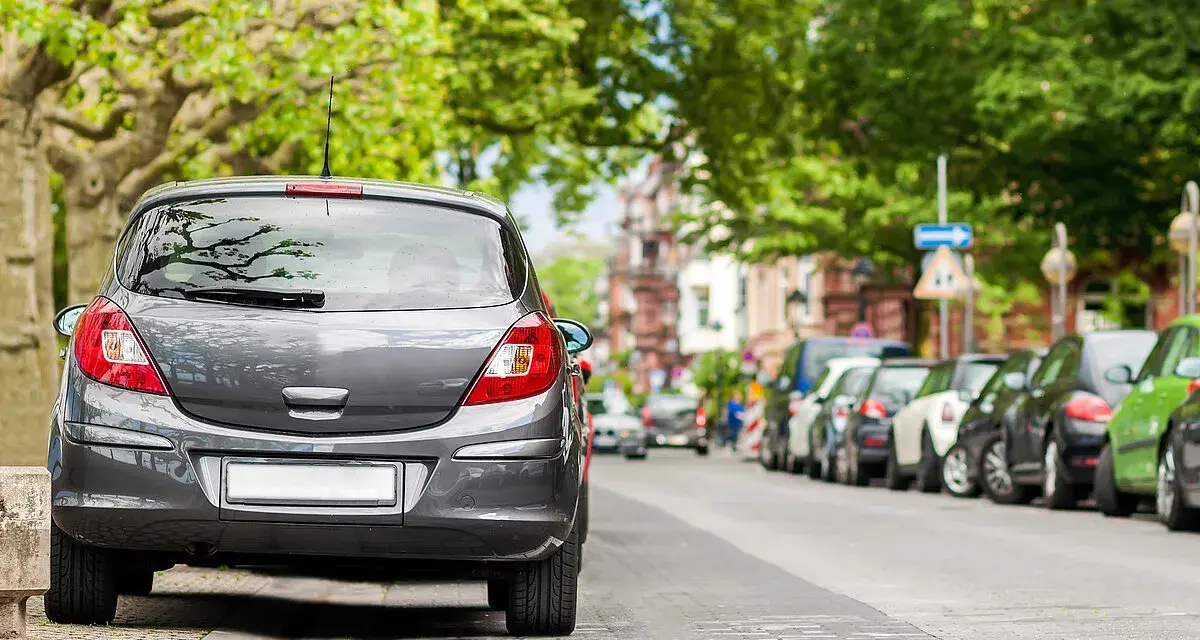 Graues Auto parkt am abgesenkten Bordstein. Mehrere Autos reihen sich die Straße entlang unter grünen Bäumen.