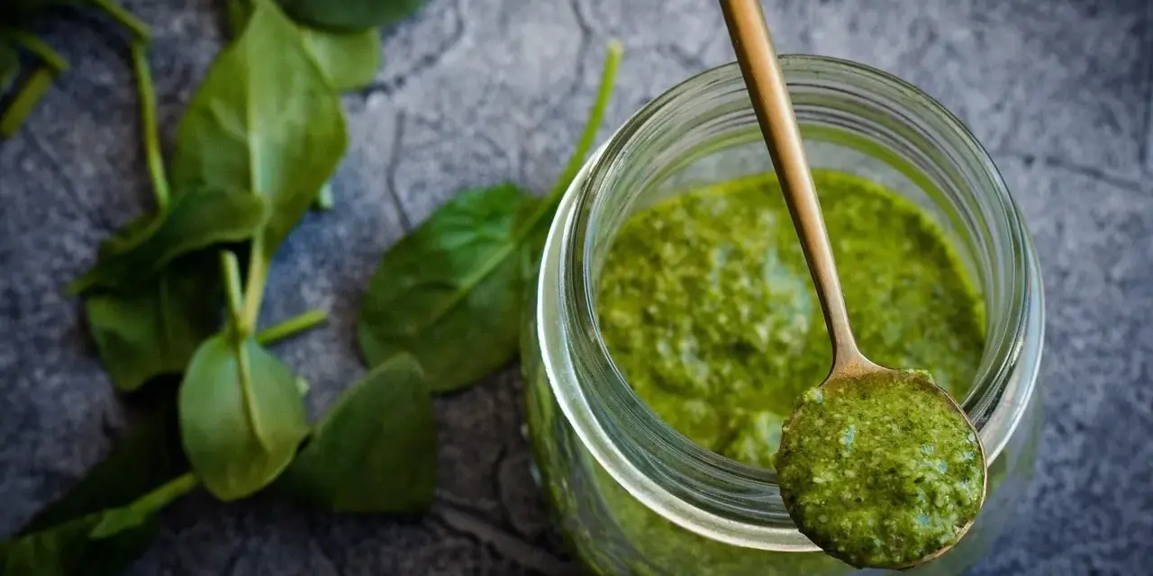 Słoik z zielonym pesto, na łyżce porcja sosu. Czy pesto ze słoika się podgrzewa? Świeże liście bazylii obok.