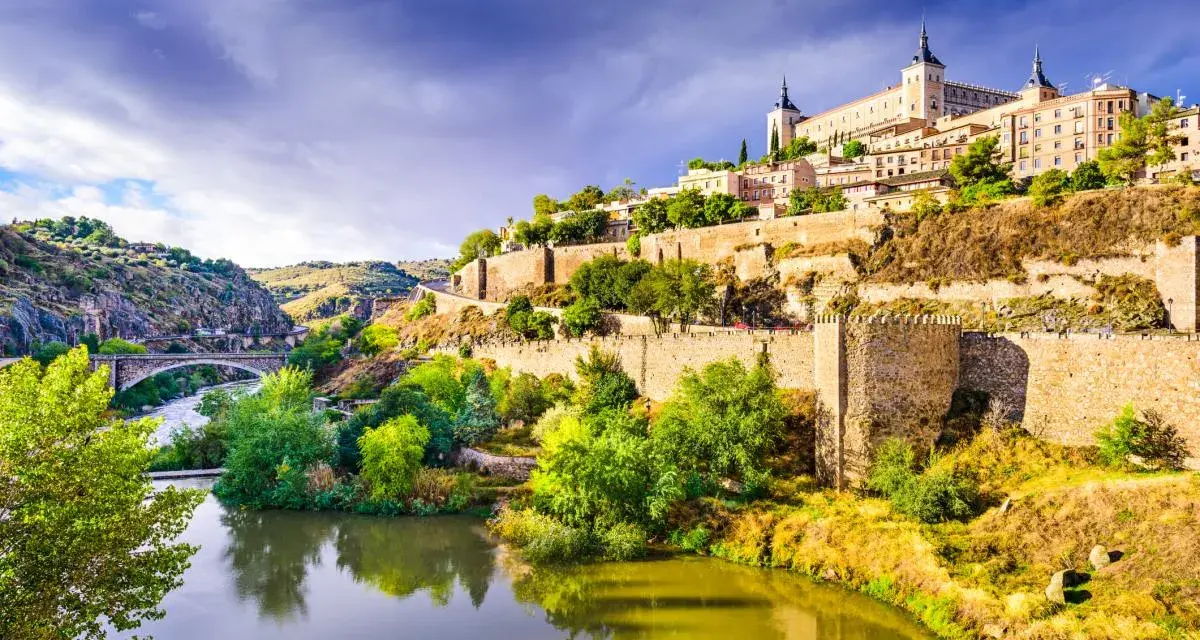 El **río Tajo** serpentea al pie de Toledo, una ciudad amurallada coronada por un imponente alcázar. Un puente de piedra cruza el agua, conectando las verdes orillas.