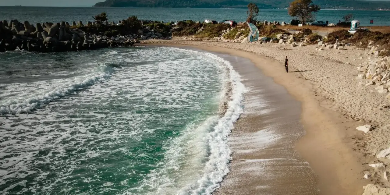 Piękne, turkusowe morze w Bułgarii obmywa piaszczystą plażę. W oddali widać wzgórza i samochody.