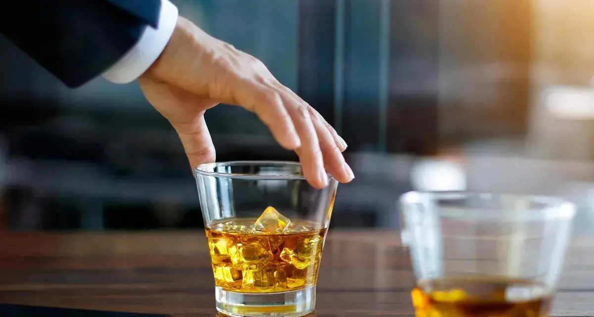 Hand greift nach einem Glas goldbraunen Whiskey mit Eiswürfeln. Ein Genuss, auch für Menschen, die glutenfreien Whiskey bevorzugen.