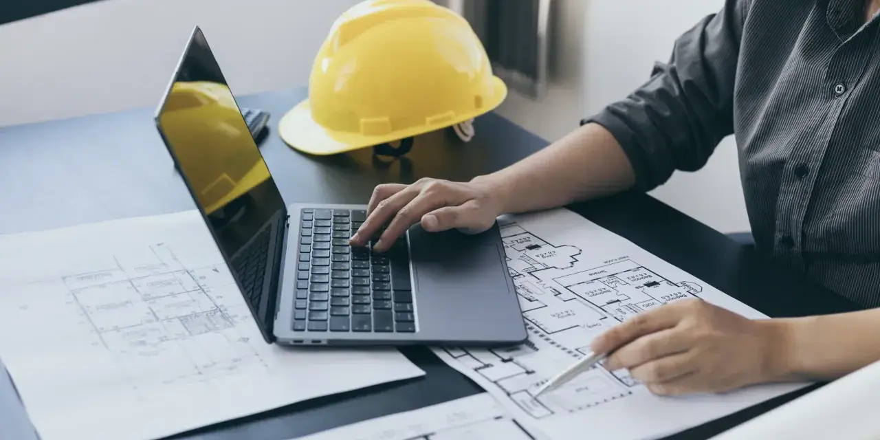 Architekt pracuje nad projektem, analizując plany i obliczając powierzchnię całkowitą budynku. Żółty kask ochronny leży obok.
