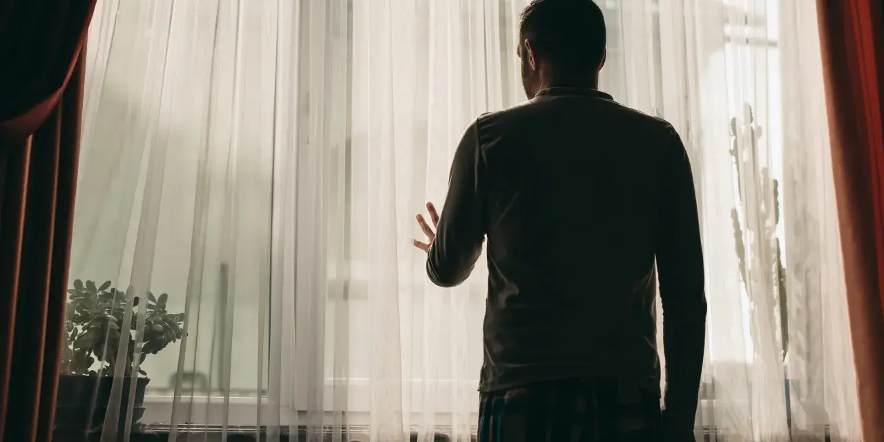 Mężczyzna stoi przy oknie, zasłoniętym przezroczystymi zasłonami. Jego dłoń opiera się o materiał, jakby szukał kontaktu ze światem zewnętrznym, co może być jednym z objawów schizofrenii.