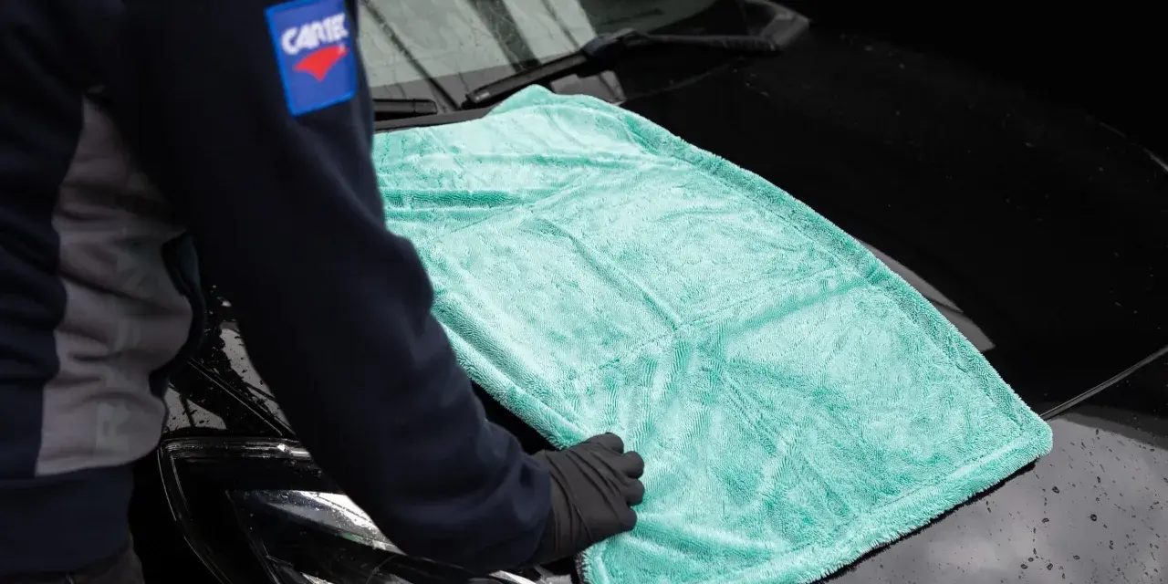 Person i handskar lägger en turkos mikrofiberduk på en svart bilhuv. Duken är från the rag company.