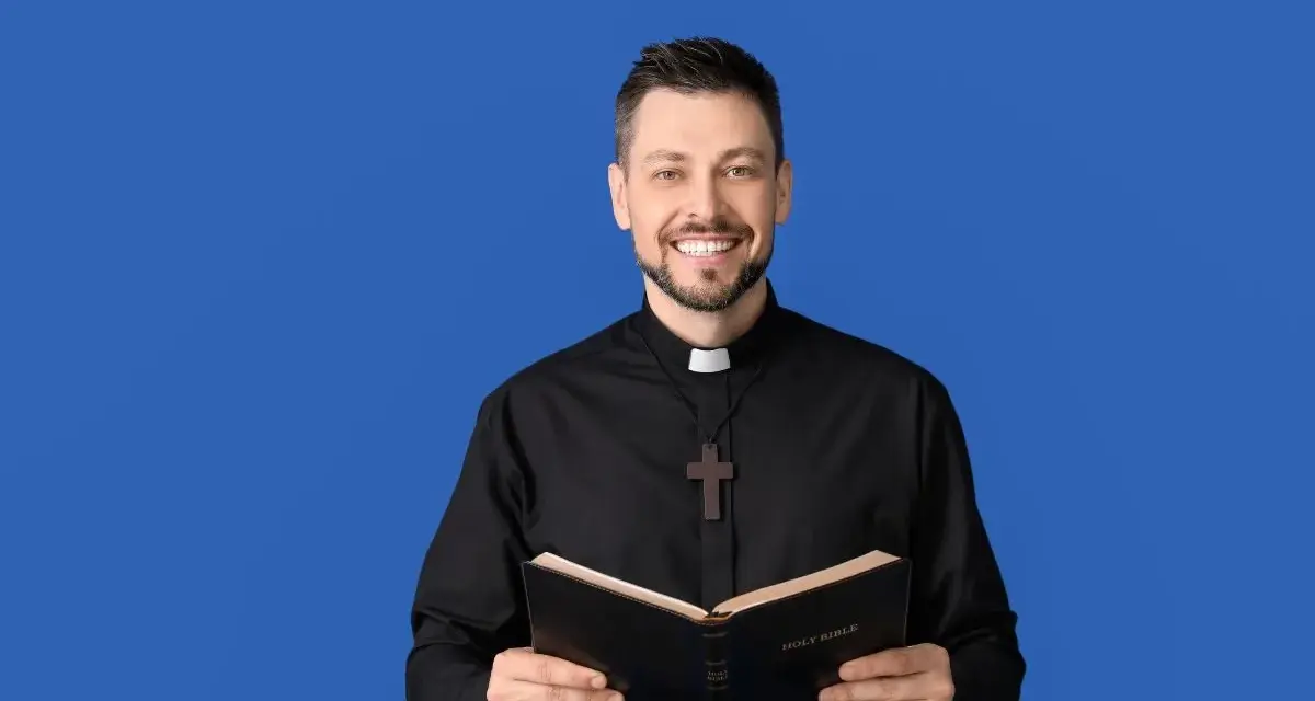 Uśmiechnięty ksiądz w czarnej sutannie z krzyżykiem trzyma otwartą Biblię. Idealny obrazek na podziękowania dla księdza.