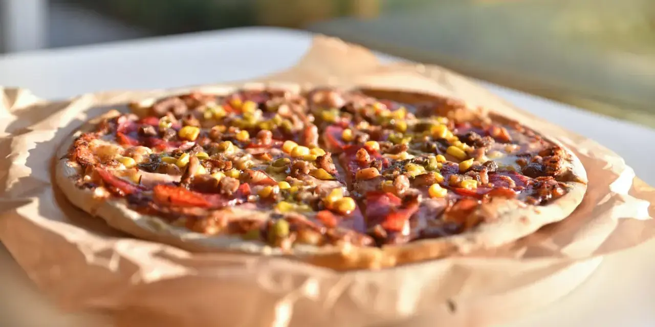 Apetyczna pizza Giuseppe 4 sery, z kukurydzą i mięsem, na papierze do pieczenia, w słonecznym świetle.