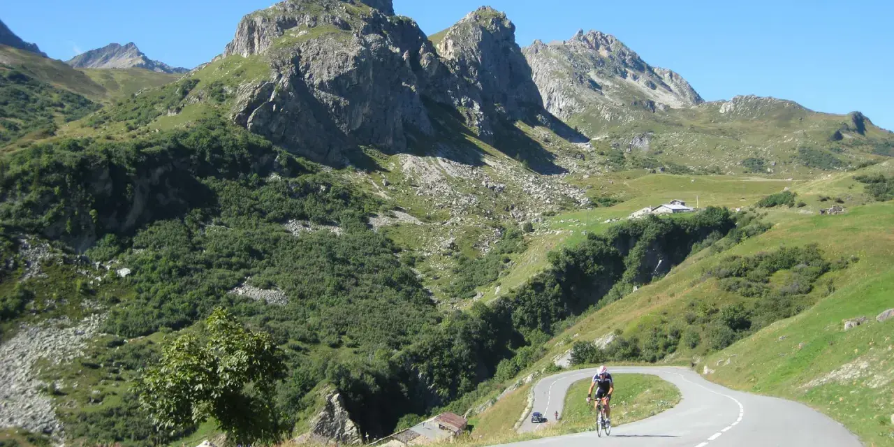 Ciclista su strada tortuosa verso il Cormet de Roselend, con montagne imponenti e verdi pendii.