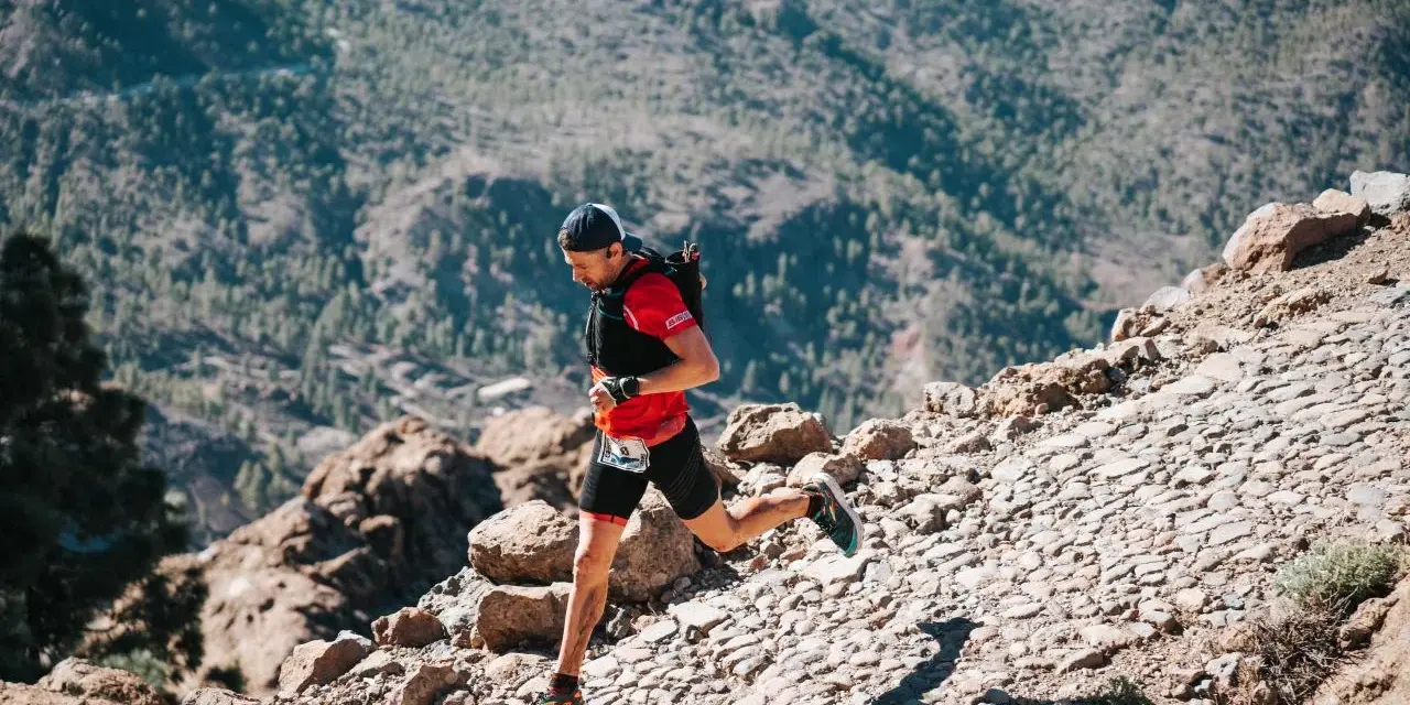 Un corredor de trail desciende por un sendero rocoso y empinado, con montañas boscosas al fondo.