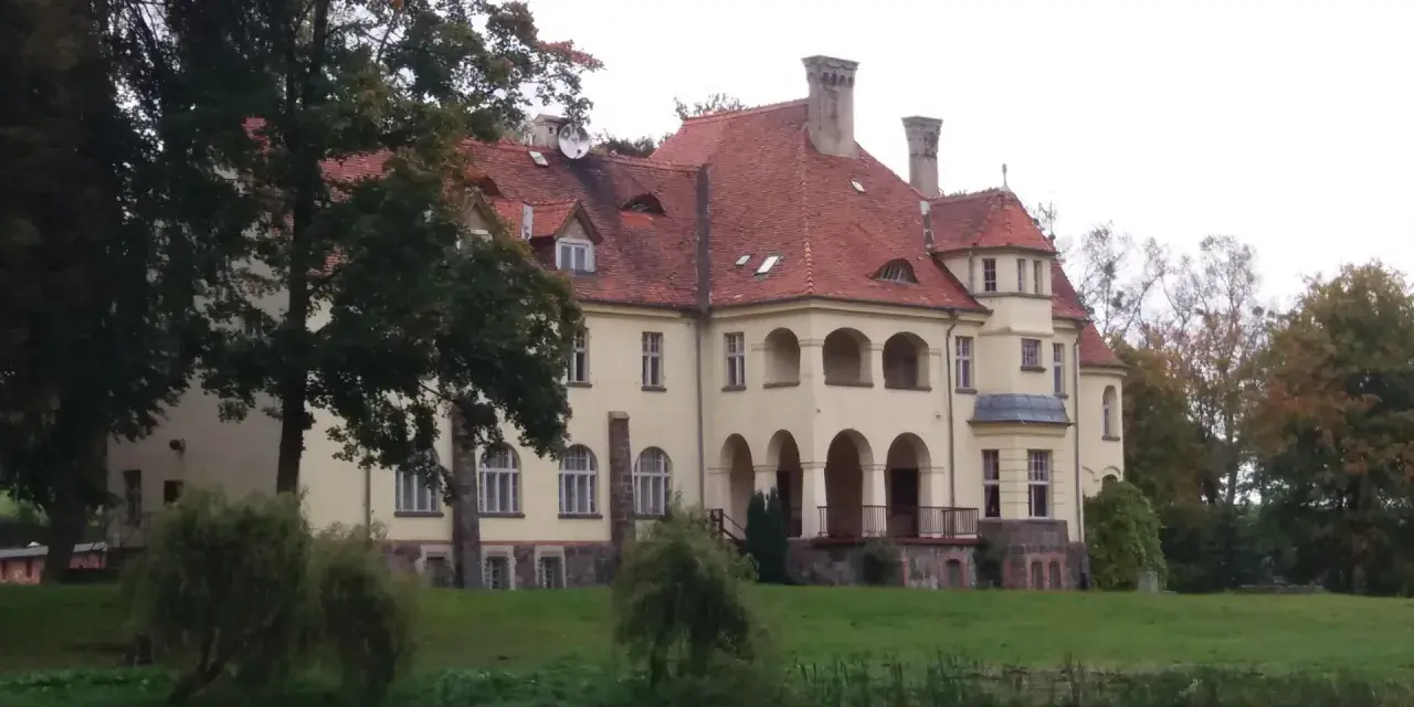 Malowniczy pałac z czerwoną, dachówkową, spadzistą dachem, otoczony zielenią i wodą.
