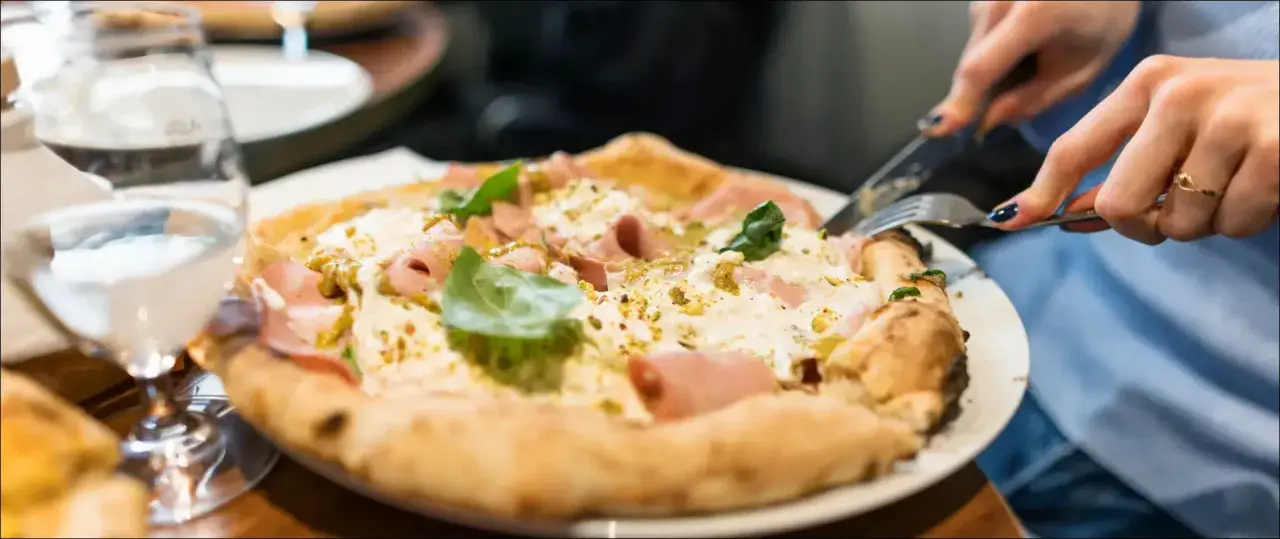 En person skär i en pizza bianco med skinka och basilika. Ett glas vatten står bredvid. Perfekt vin till pizza bianco.