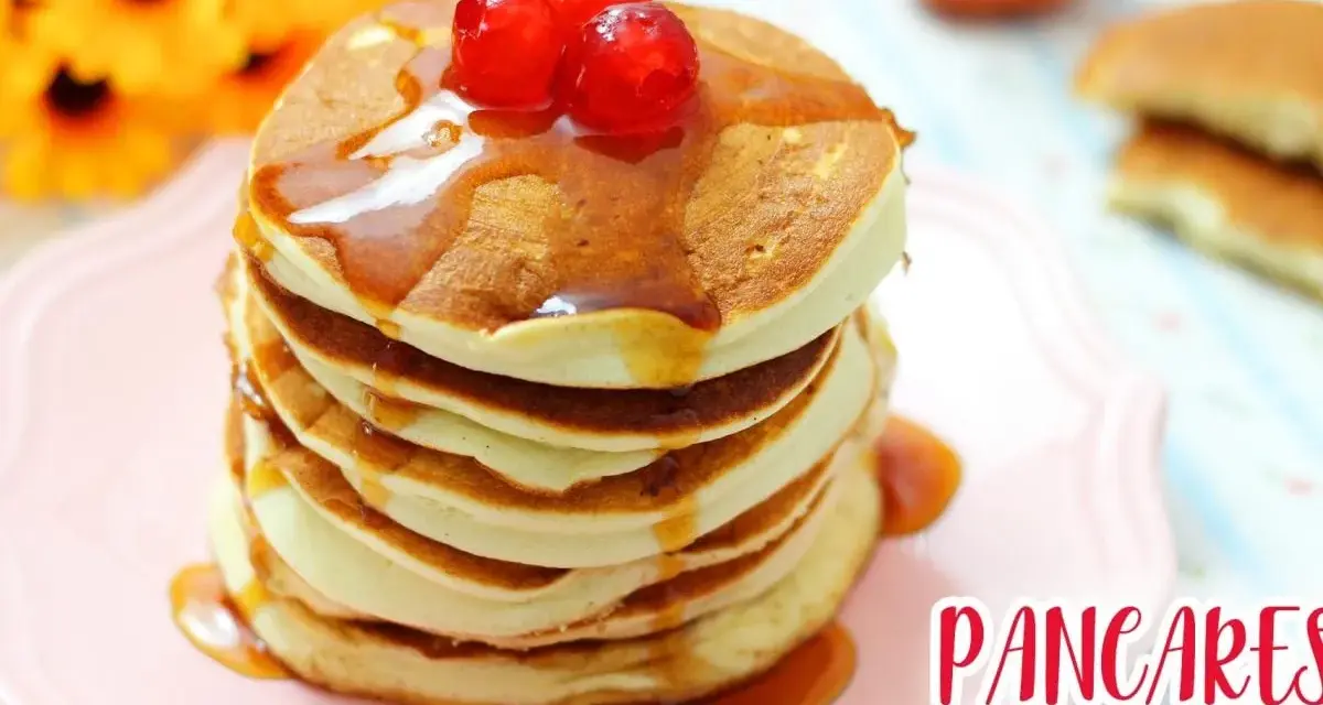 Pancakes senza glutine, soffici e dorati, impilati su un piatto rosa, con sciroppo d'acero e ciliegie rosse.