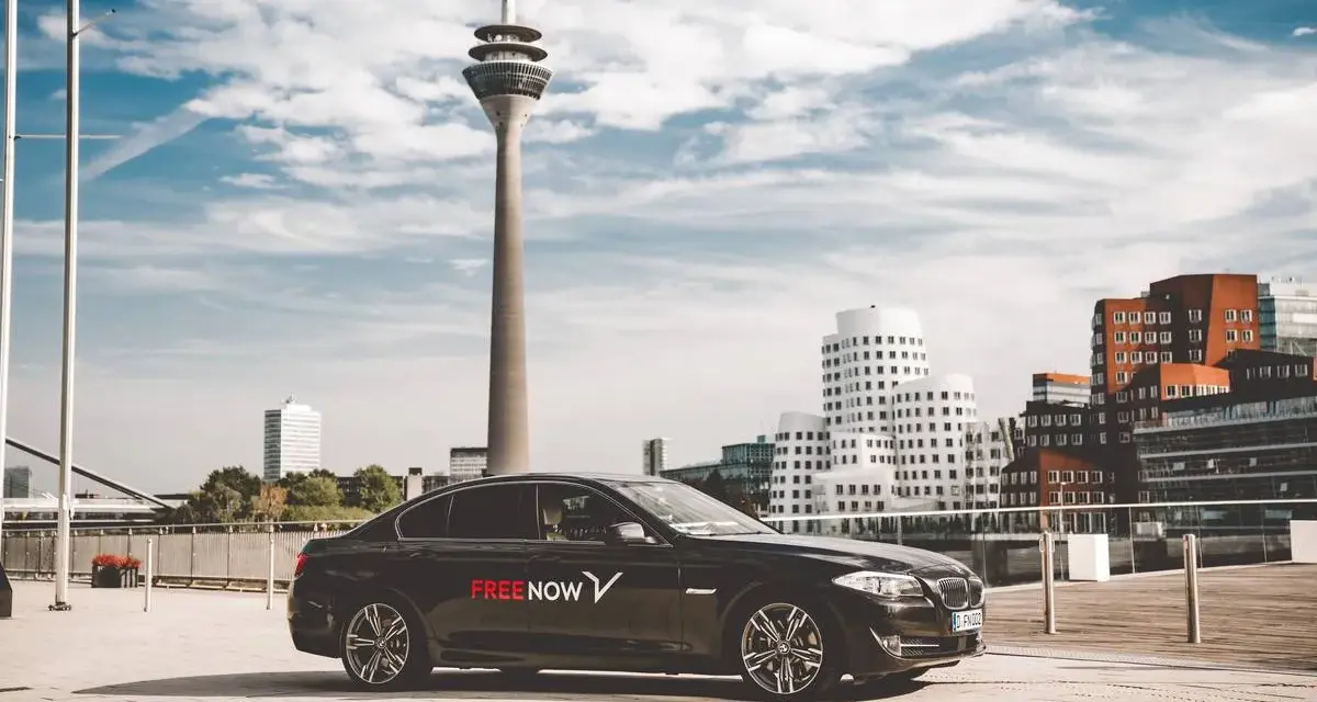 Schwarzer Wagen mit FREE NOW Logo steht vor dem Rheinturm in Düsseldorf. Ein FREE NOW Fahrer wartet auf seine Fahrgäste.