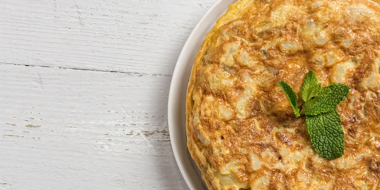 Frittata di cipolle dorata e soffice, guarnita con foglie di menta fresca, su un piatto bianco appoggiato su un tavolo di legno sbiancato.