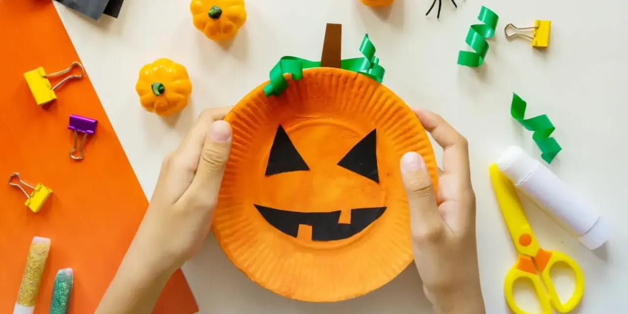 Barn gör halloween pyssel med en pumpa av papper. Små pumpor, spindlar och glitterpennor ligger redo.