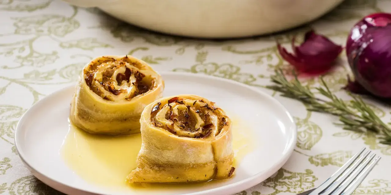 Zwei goldbraune Krautkrapfen im Sud auf einem Teller, im Hintergrund ein Auflauf mit weiteren Krapfen, Zwiebeln und Rosmarin. Ein leckeres Krautkrapfen Rezept.