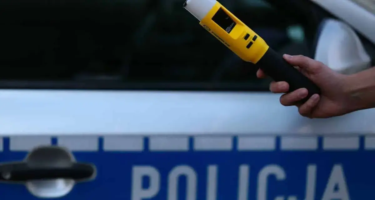 Dłoń trzymająca żółty alkomat na tle radiowozu policyjnego z napisem "POLICJA".