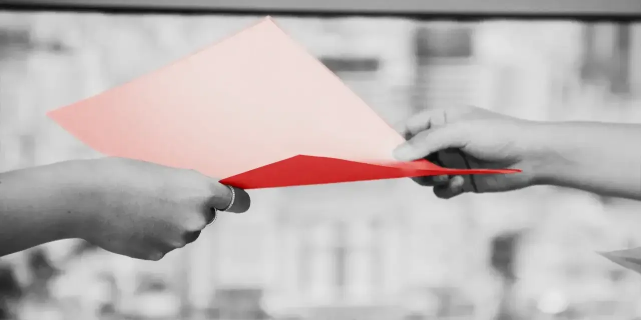 Dwie dłonie przekazują sobie dokumenty. Jedna z nich trzyma różową kartkę, druga czerwoną. Symbolizuje to uprawomocnienie postanowienia sądu.