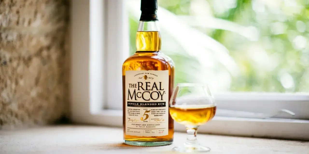 Ein besonderer Rum, "The Real McCoy", steht neben einem Glas mit bernsteinfarbener Flüssigkeit vor einem Fenster mit grünem Bokeh.