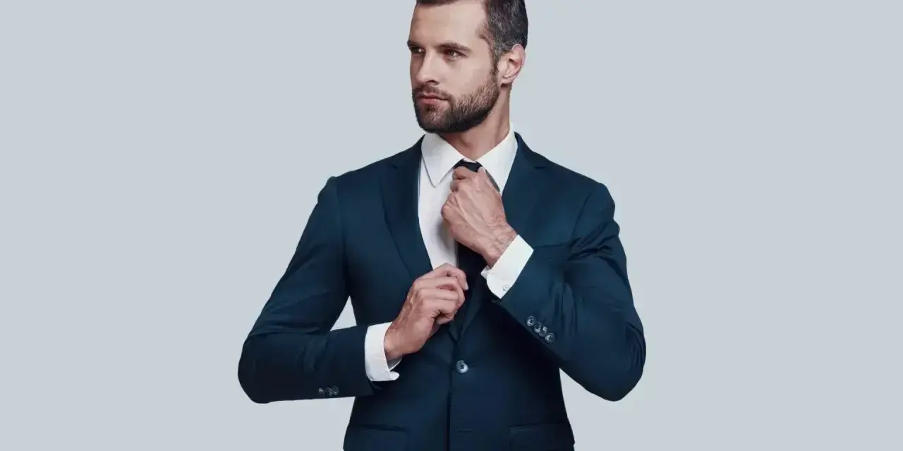 Elegancki mężczyzna w granatowym garniturze i białej koszuli poprawia krawat. Idealna stylizacja, gdy zastanawiasz się, jaka koszula do granatowego garnituru.