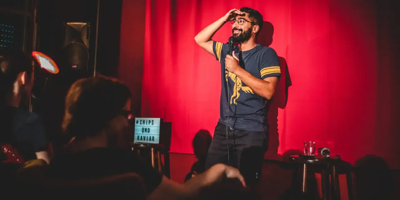 Ein Comedian steht auf einer Bühne in Berlin, hält ein Mikrofon und wischt sich die Stirn. Ein Schild mit "Chips und Kaviar" steht daneben.
