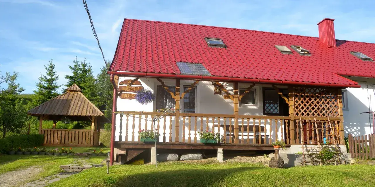 Uroczy domek z czerwoną dachem i drewnianą werandą, idealny na noclegi blisko Tarnicy. Obok altana i zielony ogród.