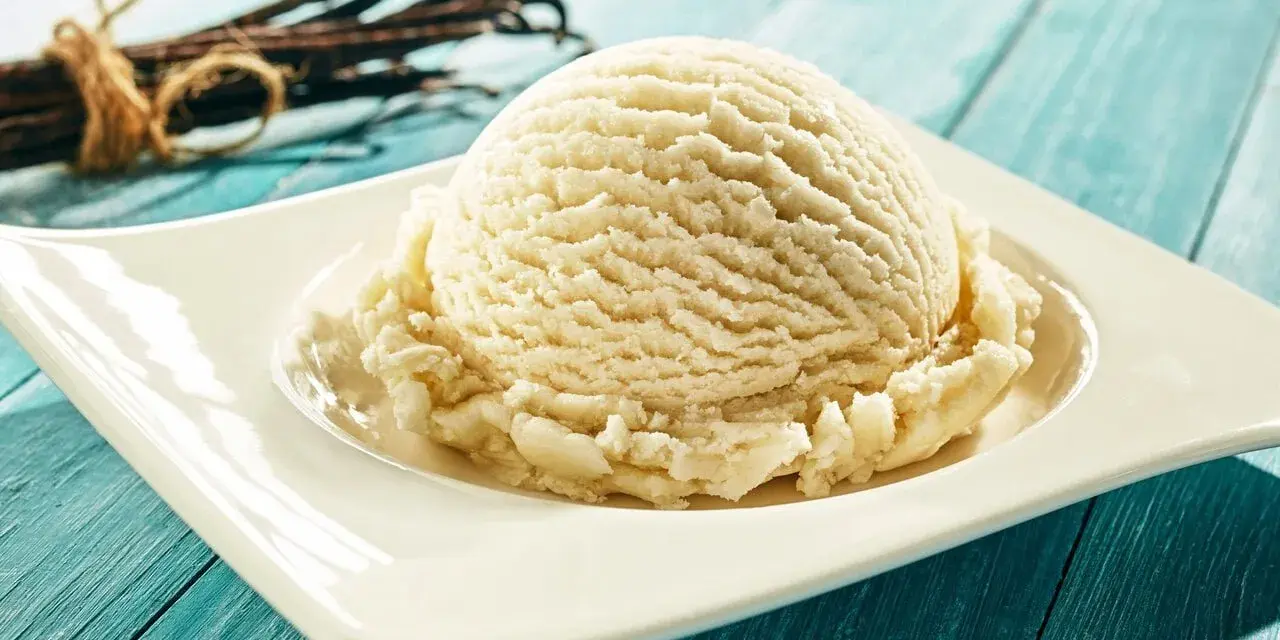Une boule de glace maison à la vanille, servie sur une assiette blanche, avec des gousses de vanille en arrière-plan.