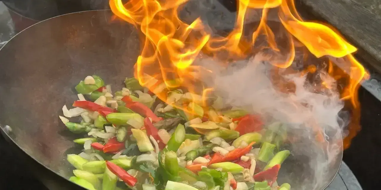 Warzywa w woku płoną w ogniu, tworząc aromatyczny stir fry. W tle widać drugie danie.