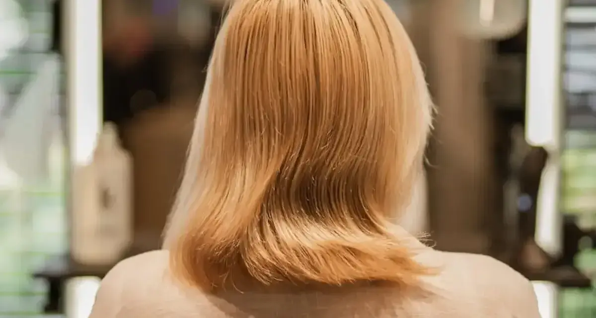 Blond włosy po zabiegu farba demi permanentna, delikatnie pofalowane, w tle rozmyte światła salonu fryzjerskiego.