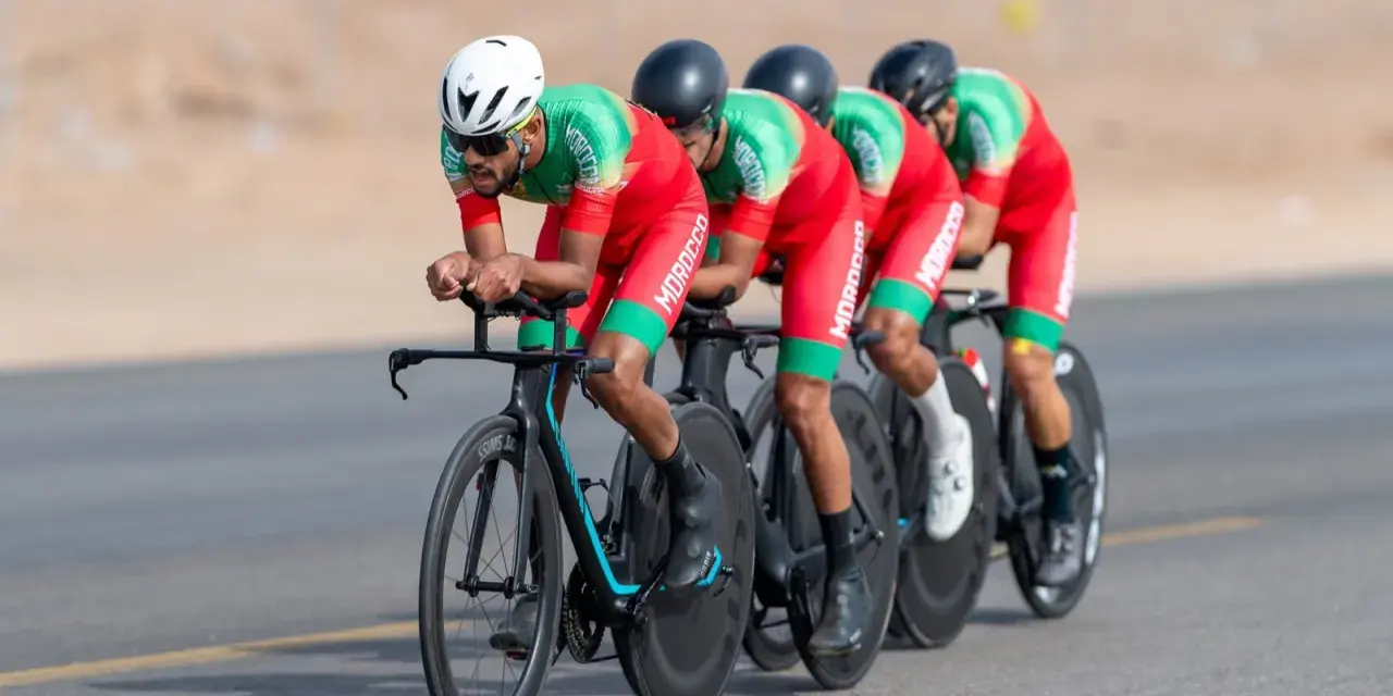 Équipe cycliste marocaine en tenue rouge et verte, concentrée sur la course. Ils roulent en peloton, tels des pur sang arabes noirs, sur un parcours désertique.