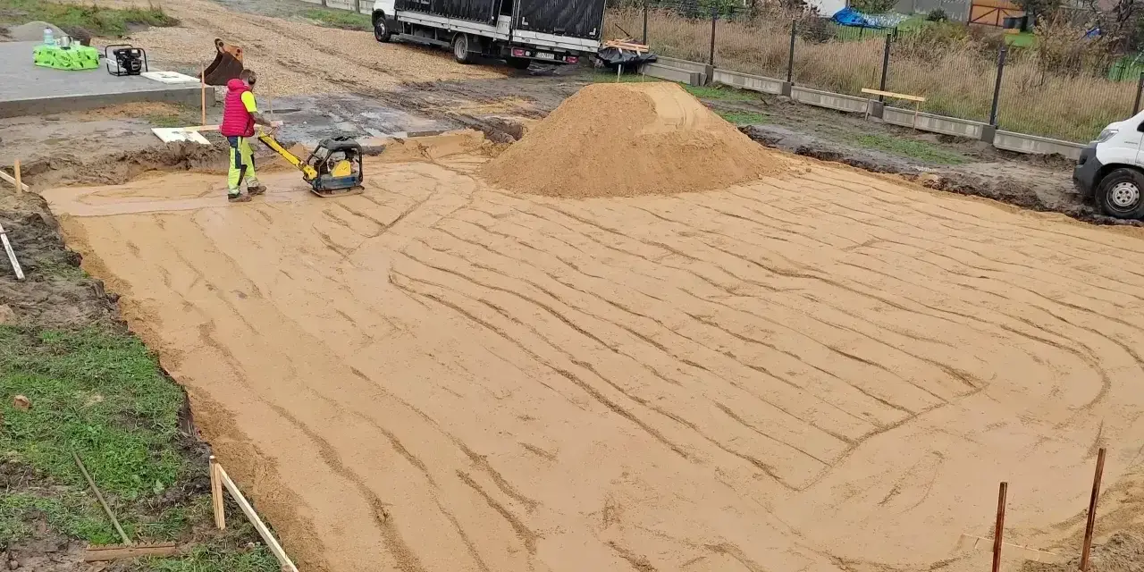Pracownik zagęszcza piasek zagęszczarką. To kluczowy etap wymiany gruntu pod fundament, zapewniający stabilność przyszłego budynku.