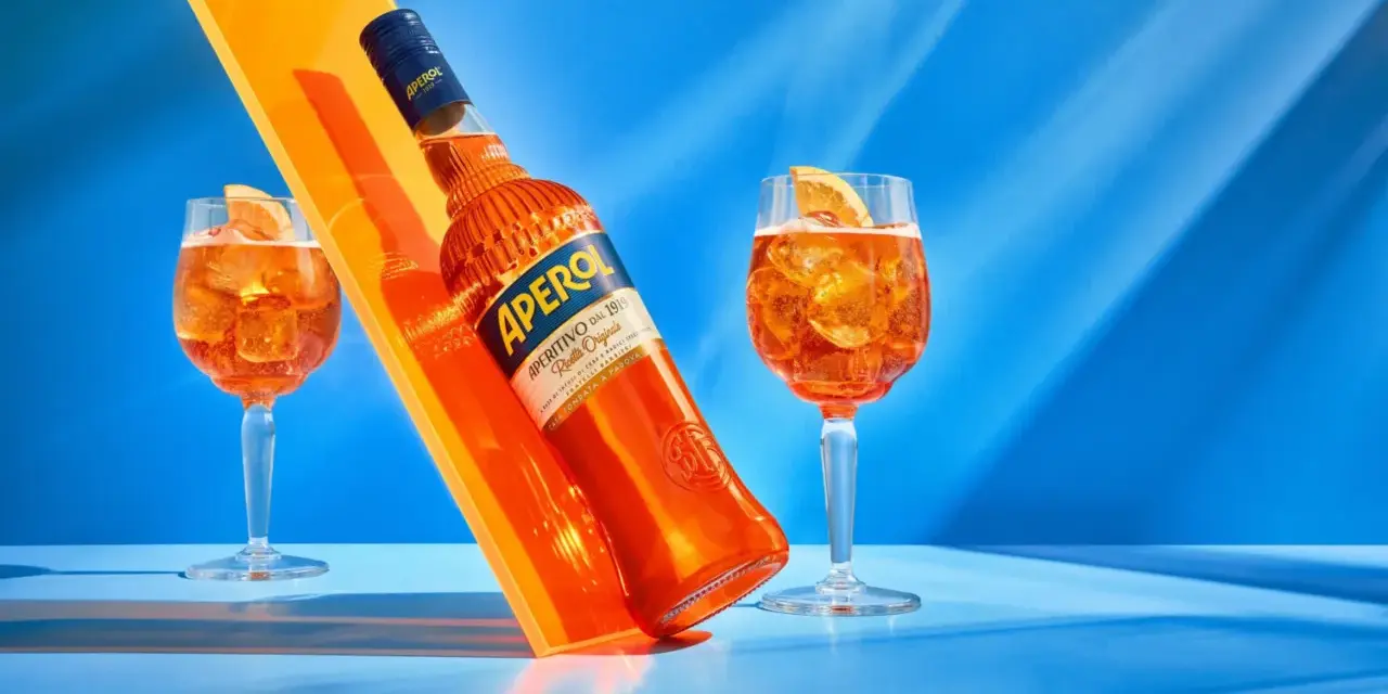 Aperol, znany z orzeźwiającego smaku, ma 11% alkoholu. Na zdjęciu butelka i dwa drinki z pomarańczą.